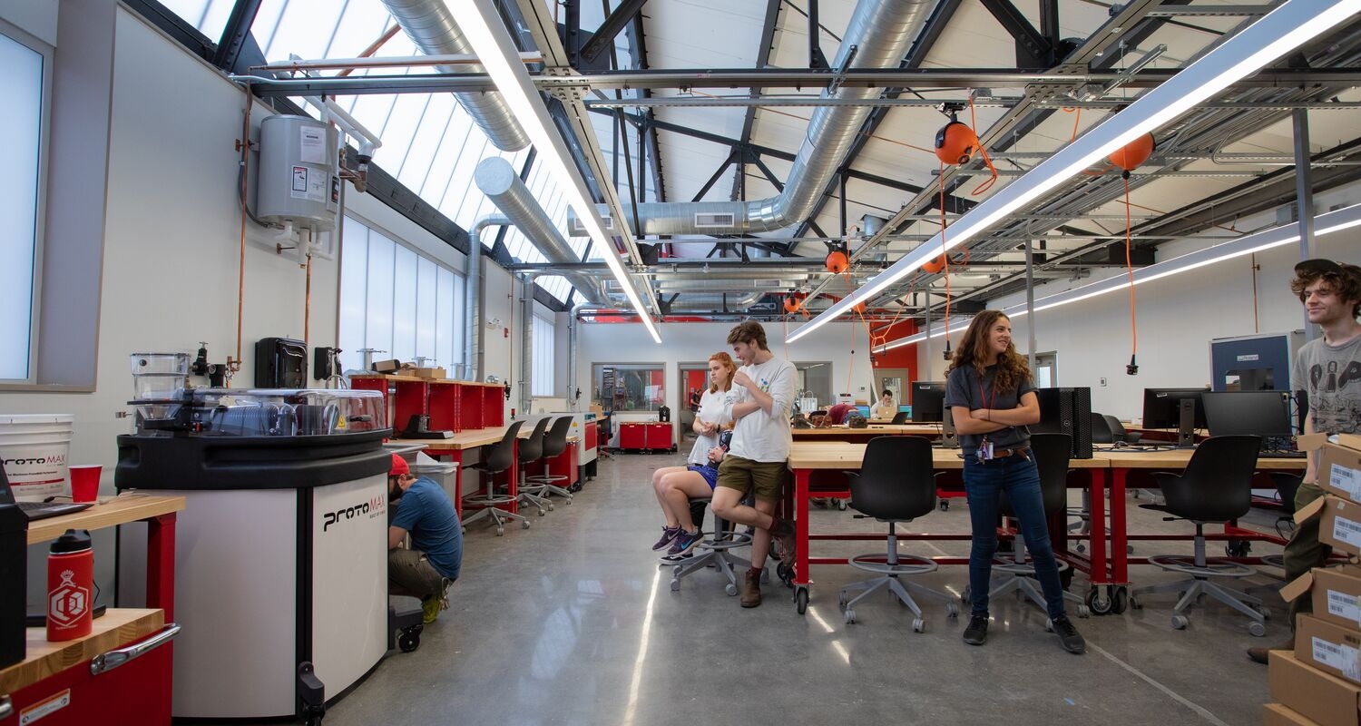 Students are gathered at a worktable in the center of a modern, sun-filled workshop space. There are exposed ducts and beams in the roof and the floor is finished concrete.