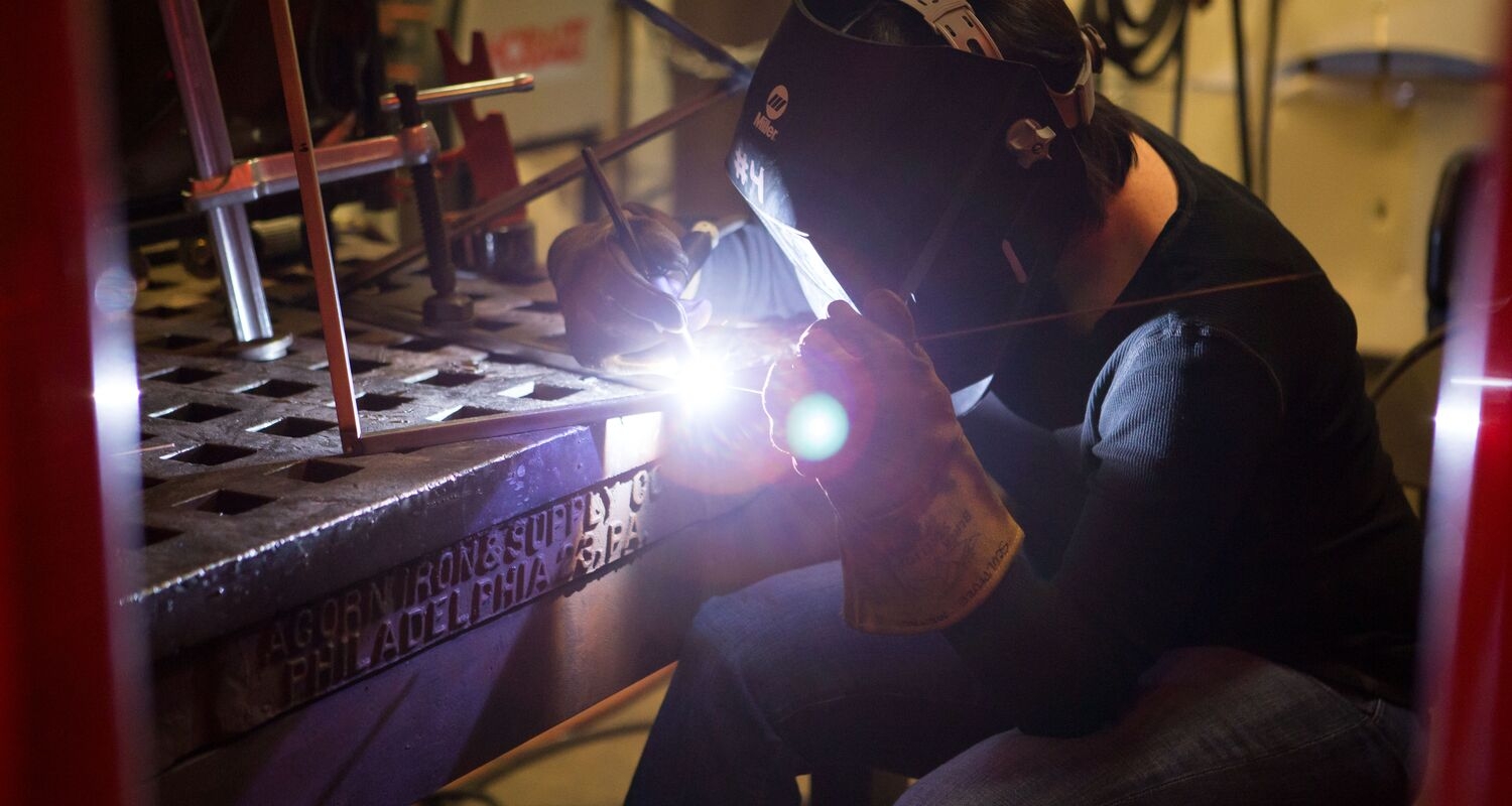 A student in a sculpture studio wearing a welding mask, using welding equipment. 