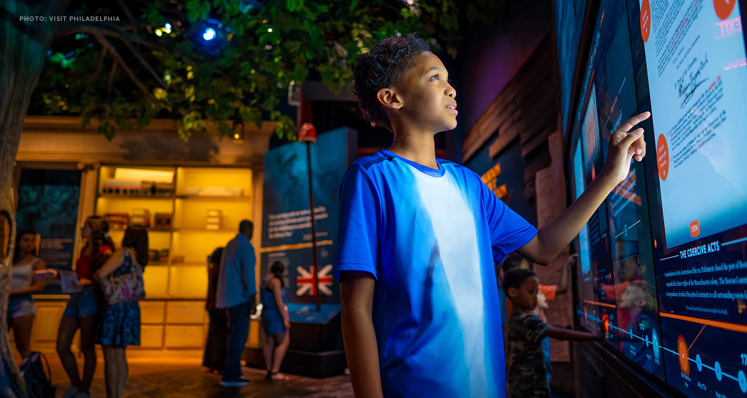 A young boy is wearing a blue shirt standing next to an interactive exhibit. Photo credit: Visit Philadelphia