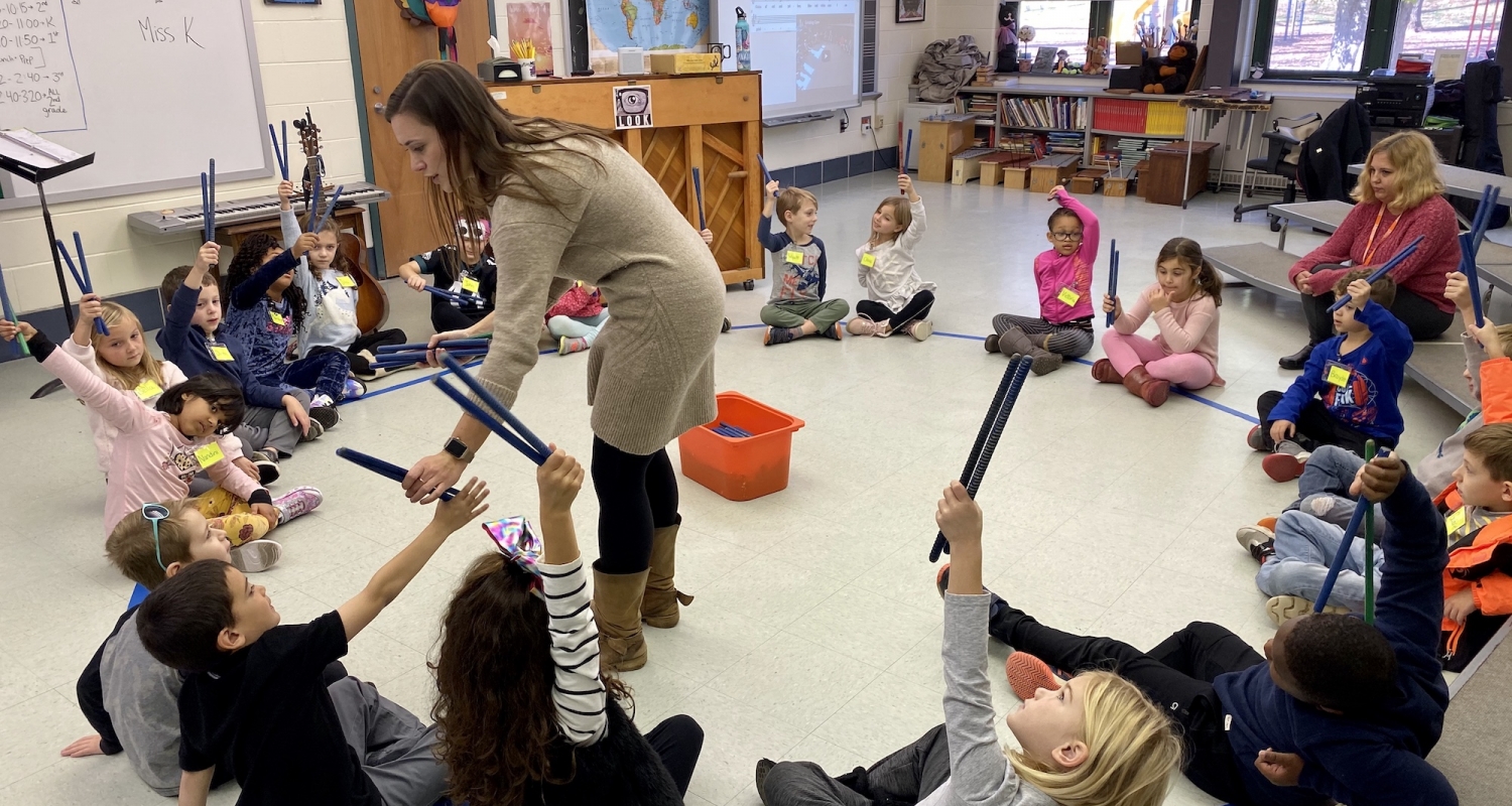 A music teacher collects rhythm sticks from students in a classroom.