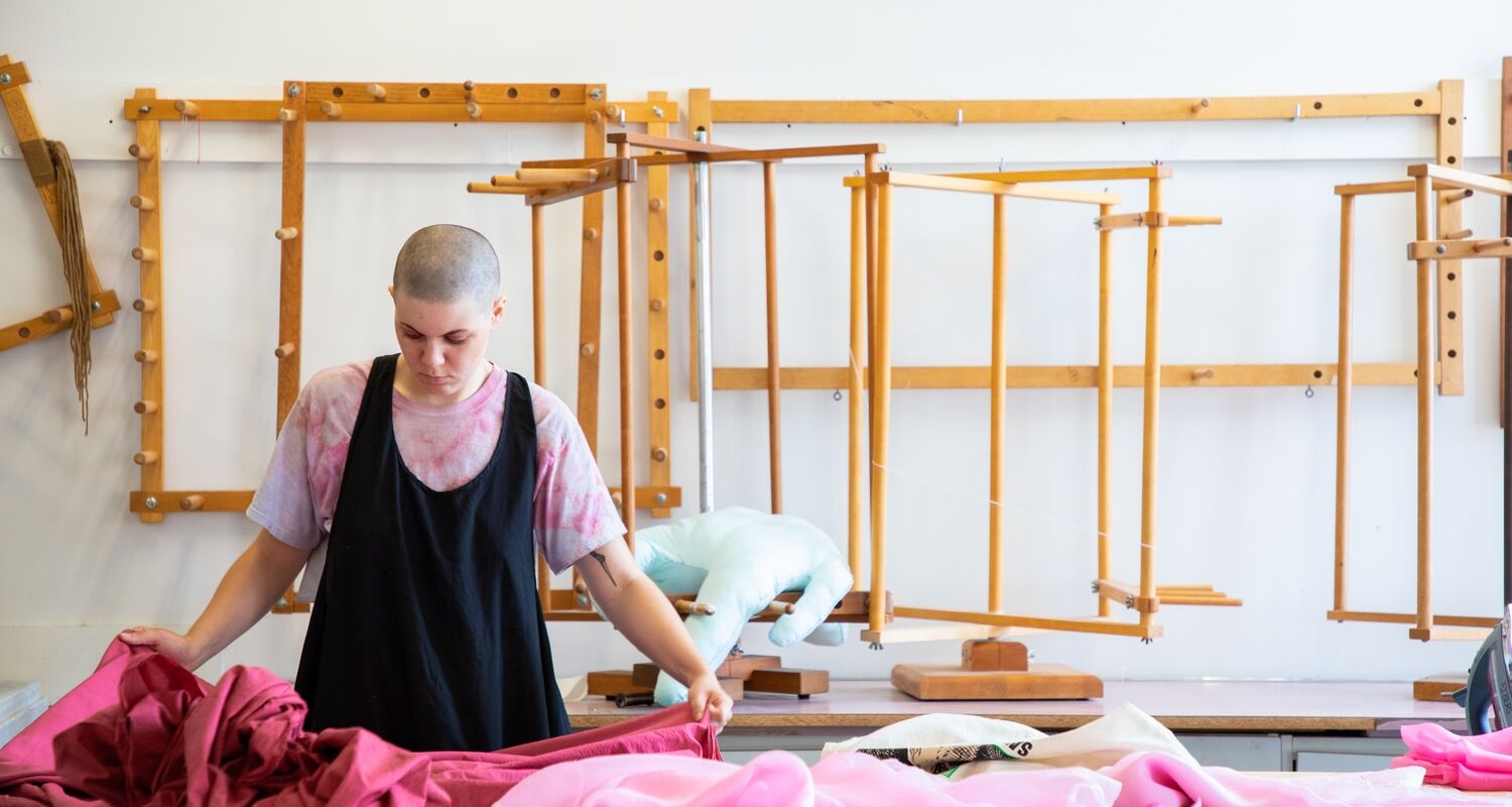 A student in the fibers studio, looking at a large sheet of pin fabric. Fiber equipment can be seen in the background.