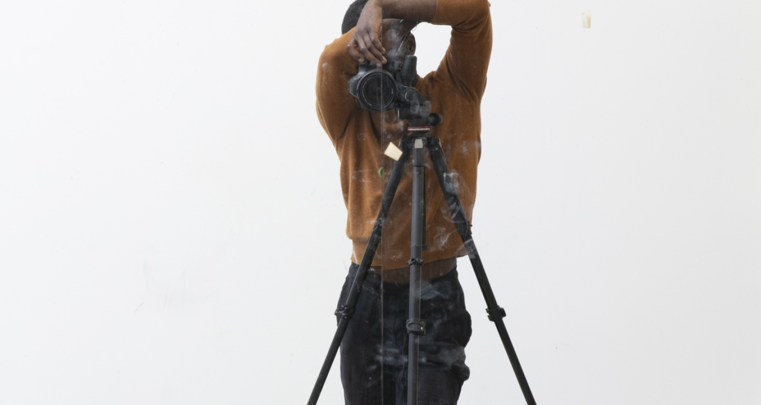 Self portrait of the artist Paul Mpagi Sepuya, artist posing in a white space behind a photo tripod with face obscured by camera