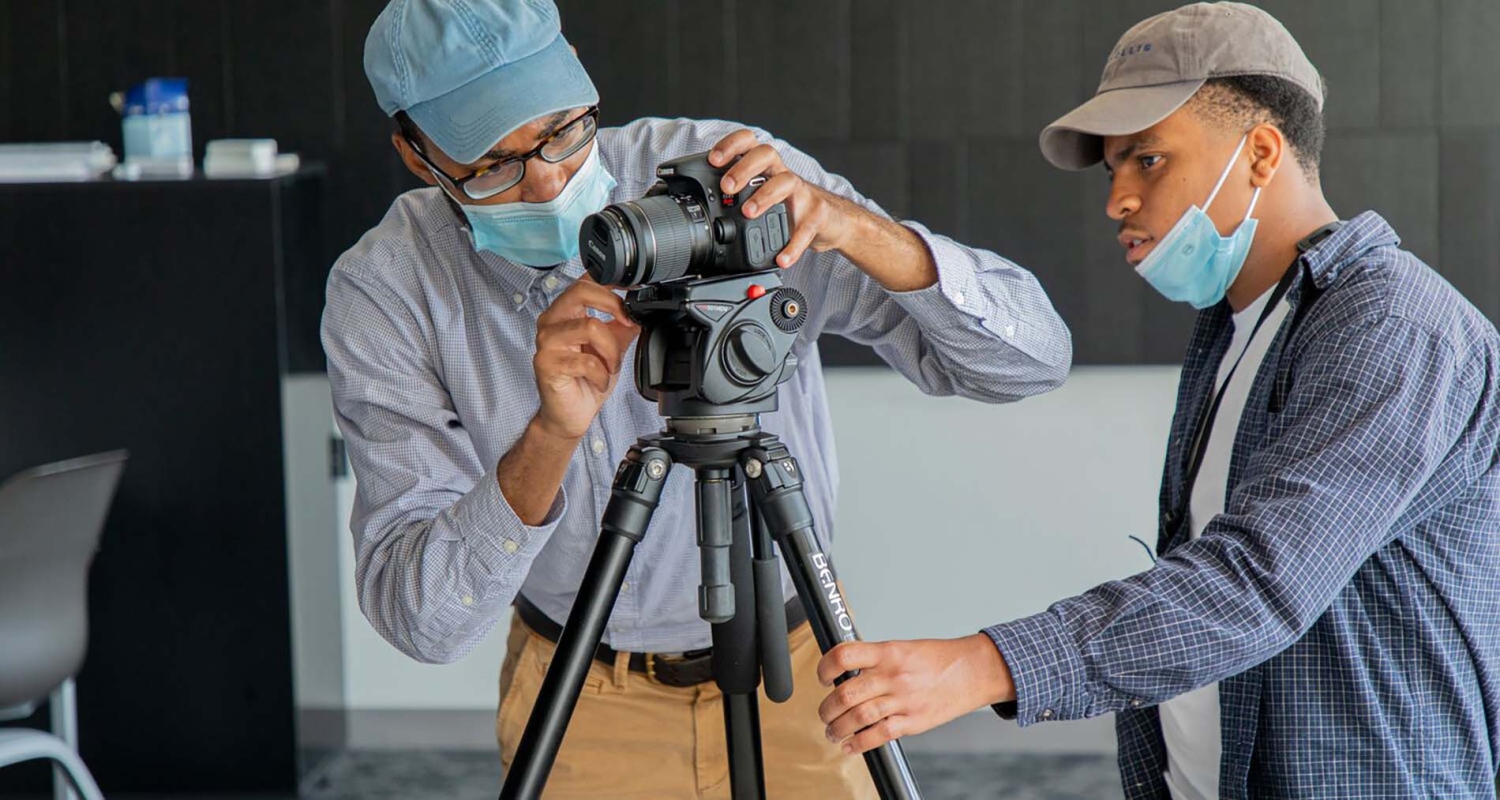 A student filmmaker in a blue button down shirt and a gray baseball hat sets up a camera shot with a faculty member in a blue shirt and tan pants and a denim baseball hat 