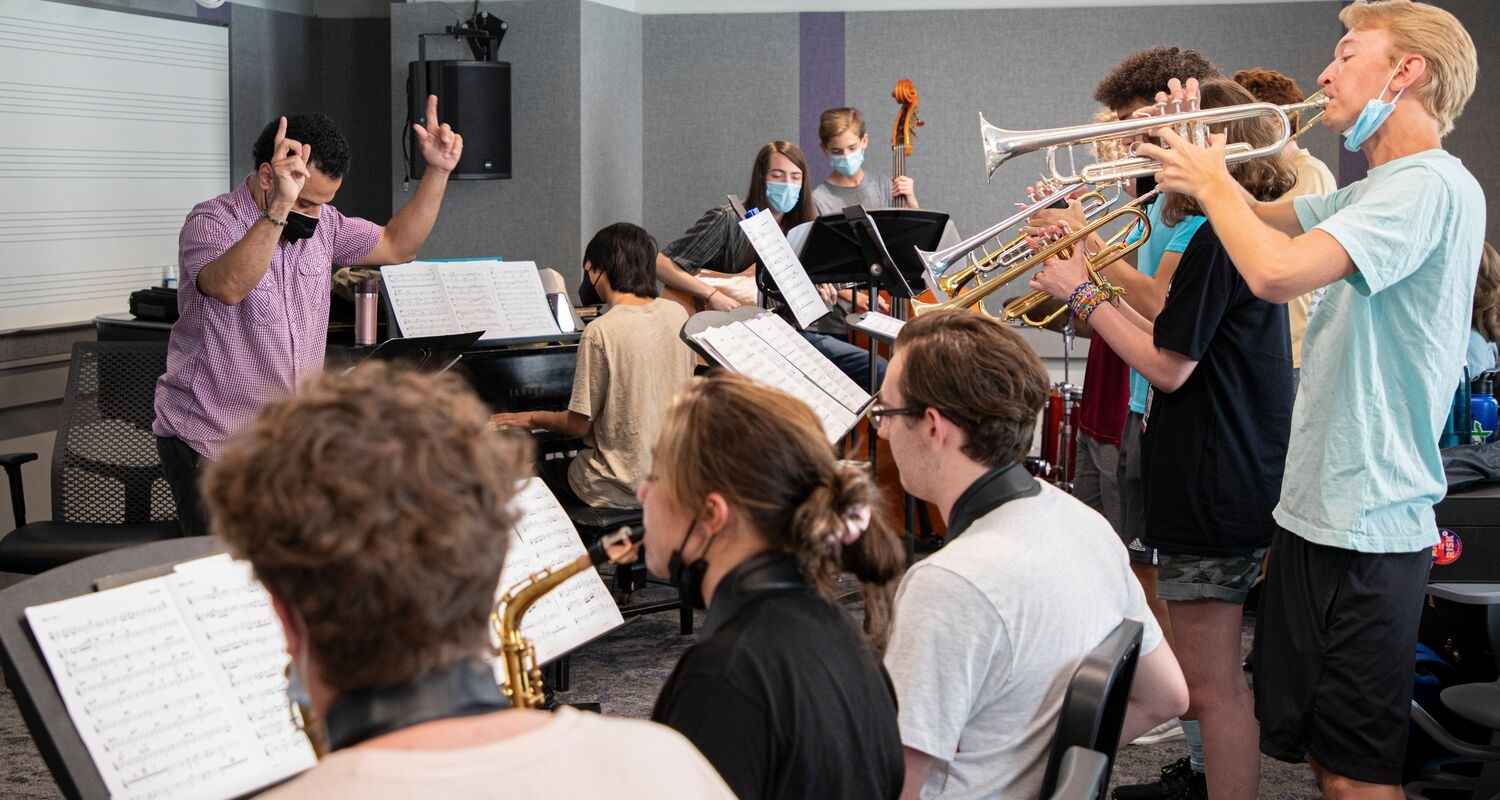 A faculty member in a purple shirt conducts an ensemble of sitting and standing student musicians who are playing trumpets and saxophones with an upright bassist in the background