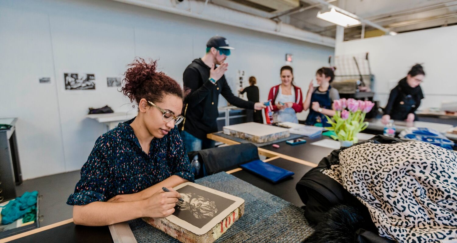 In a brightly lit printmaking studio, a student with reddish hair and glasses is working on a piece in the foreground. In the background, several students are interacting and there is a bouquet of flowers in the middle of the table they are gathered around.