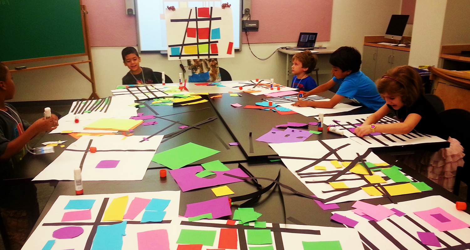 A group of elementary students sit around a large table covered in cut, multicolored construction paper. 