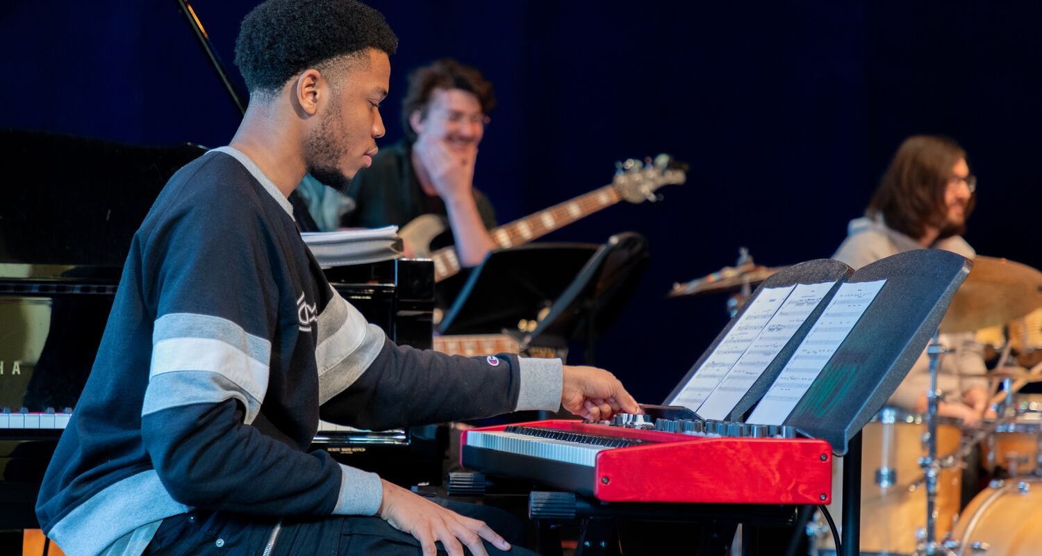 A student (a member of the Transfusion ensemble) sits at a keyboard in the foreground, his left hand is adjusting a knob. Two other students in the ensemble are visible in the background, one appears to be holding an electric guitar and the other is seated at a drum kit.