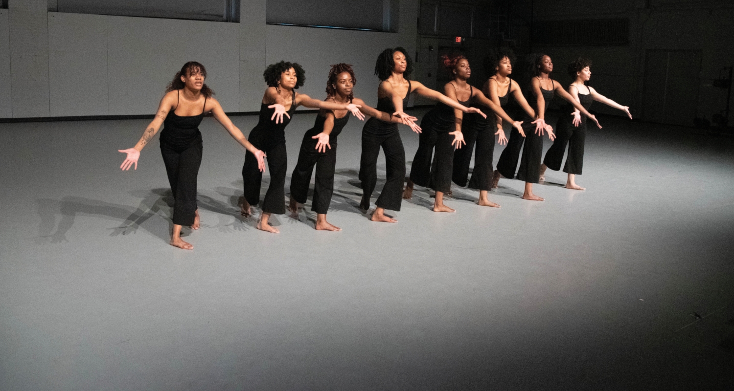 a row of dancers are in a row in a gray room, illuminated by a long-throw spotlight. all have their arms outstretched with palms upwars and are wearing matching black outfits