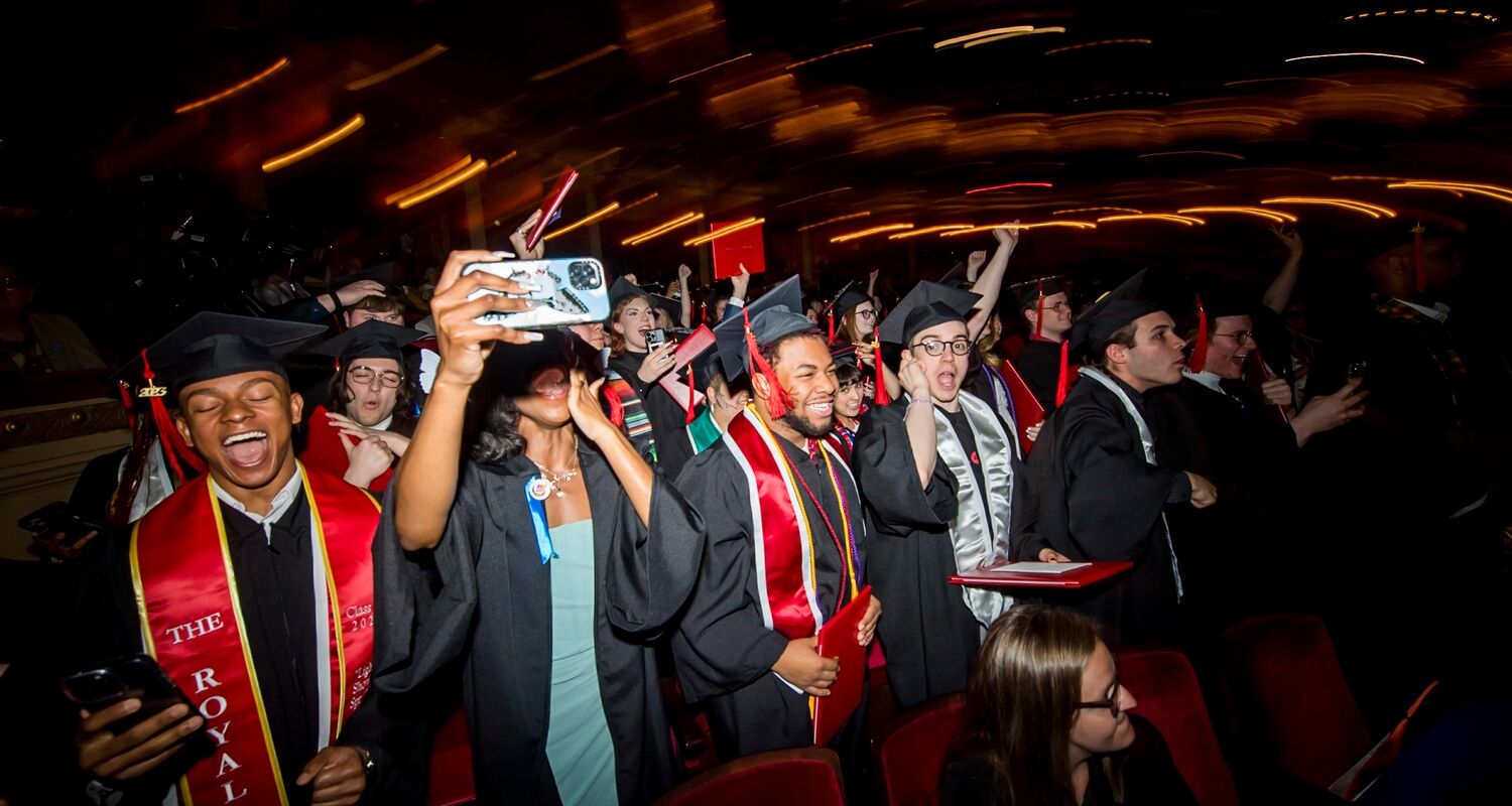 commencement 2023 in action, with rows of seated graduating students in caps and gowns. all are excited, exuberant, animated, many smiling and laughing and holding phones. the foreground is illuminated with a bright camera flash while the background is dark and blurry, streaked with warm light trails of an auditorium. 