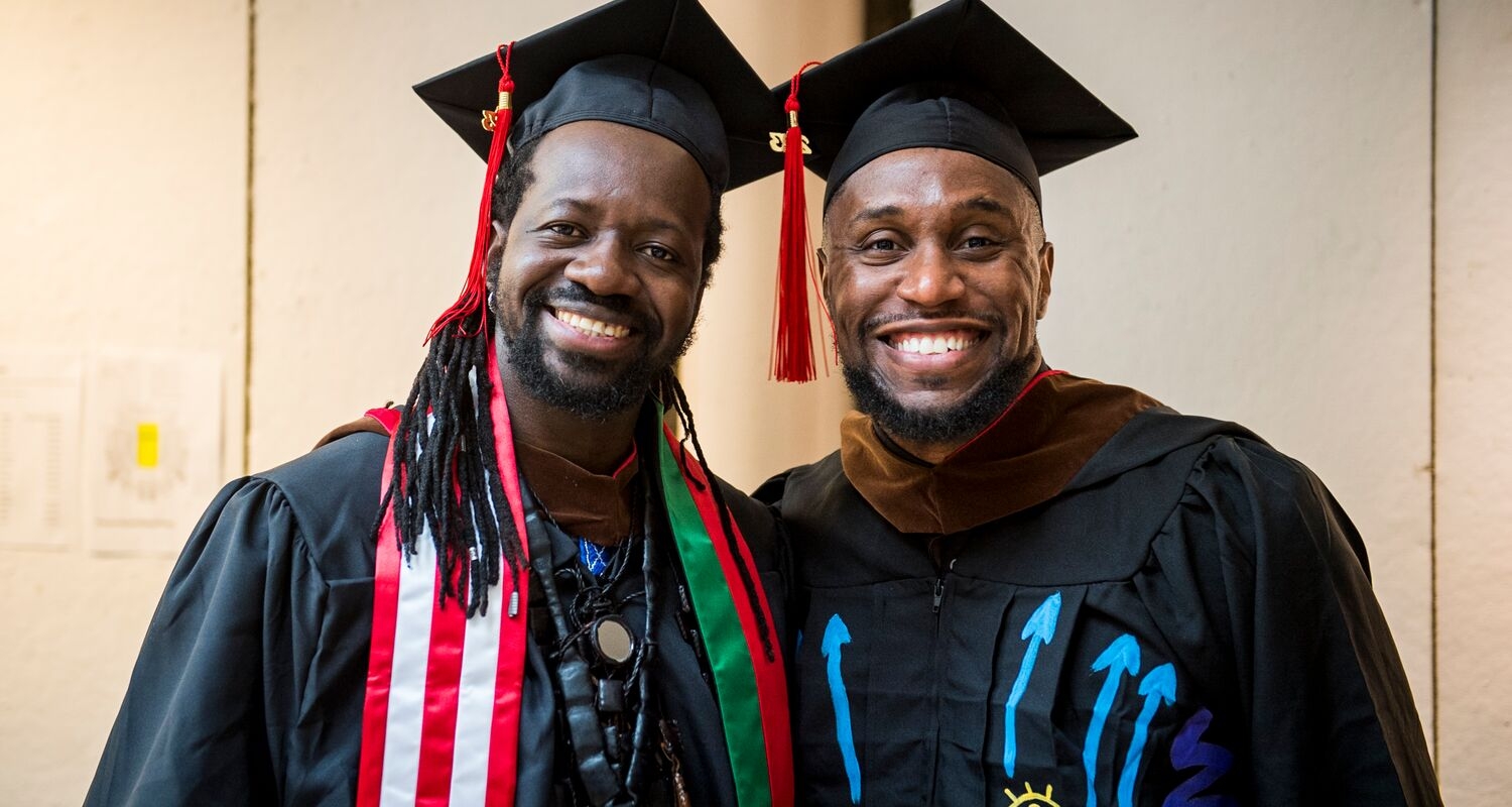 Two masters students in their caps and gowns stand in front of a tan wall and smile