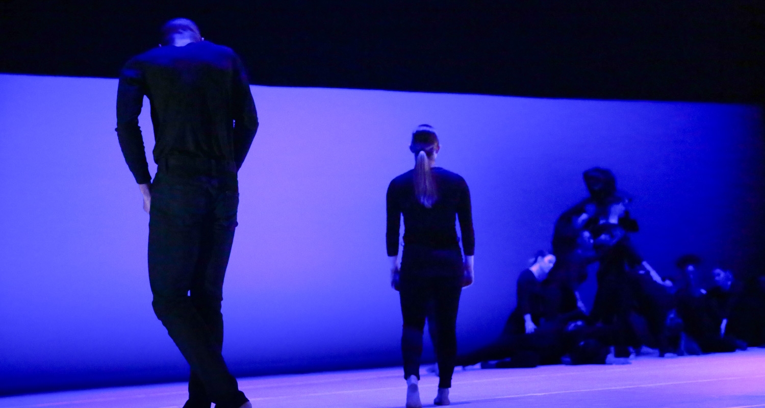Dance students perform on a stage in black costumes against a blue and purple backdrop