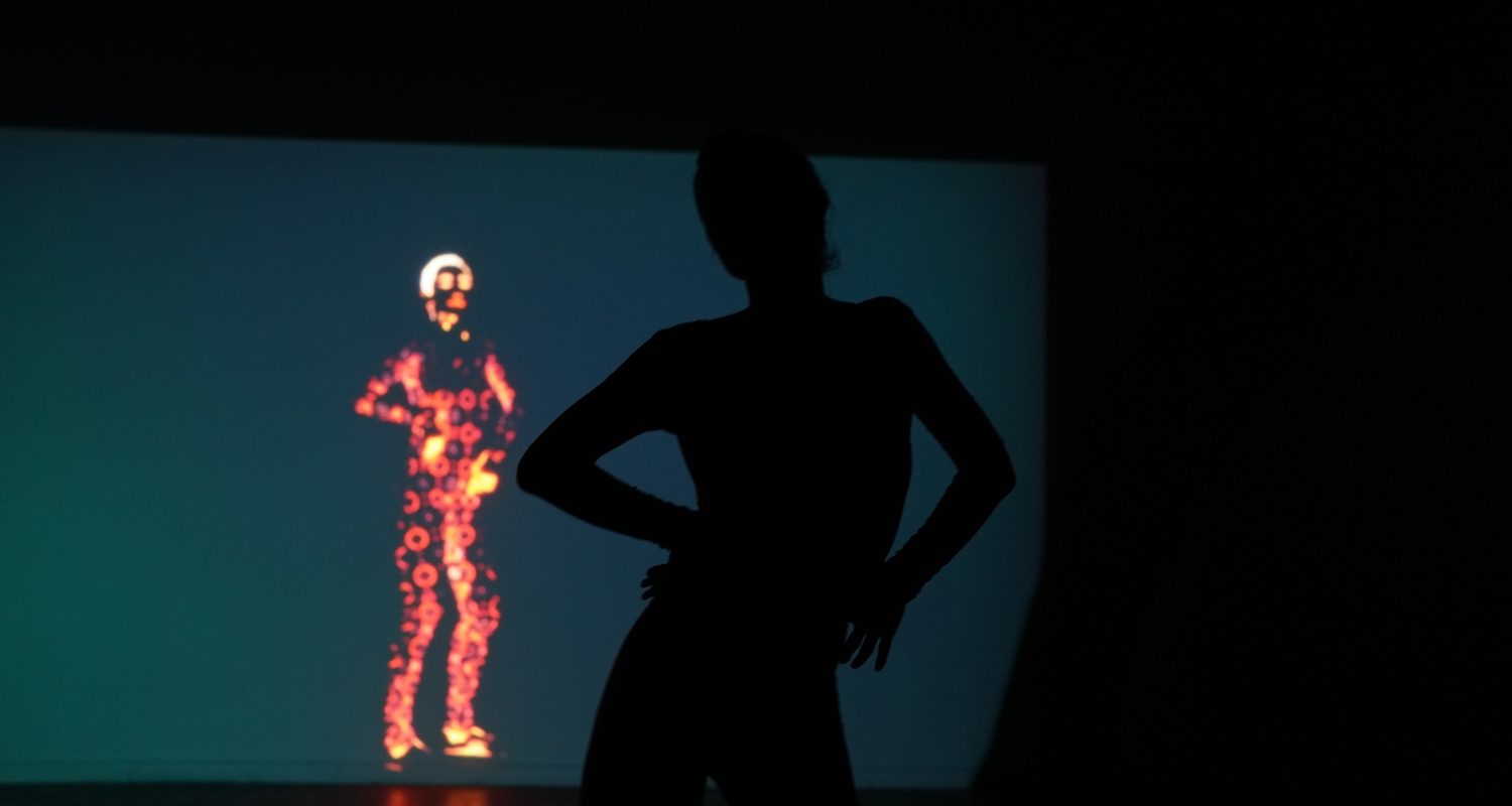 a dance performance in a dark room depicting the silhouette of a person with hands on hips standing in a dark room ahead of a wall-wide projection depicting a person rendered in glowing circular and flower patterns.  