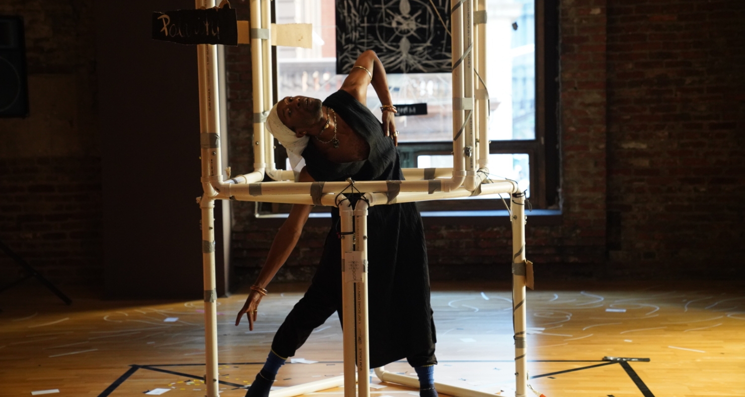 a person dancing within a sculpture consisting of PVC piping assembled into two stack by rotationally offset cubes. the sculpture is in the center of a black lined rectangular diagram on the wooden floor within a dark room with a large window. the person is wearing a black smock or tunit and has one hand stretched to the floor. 