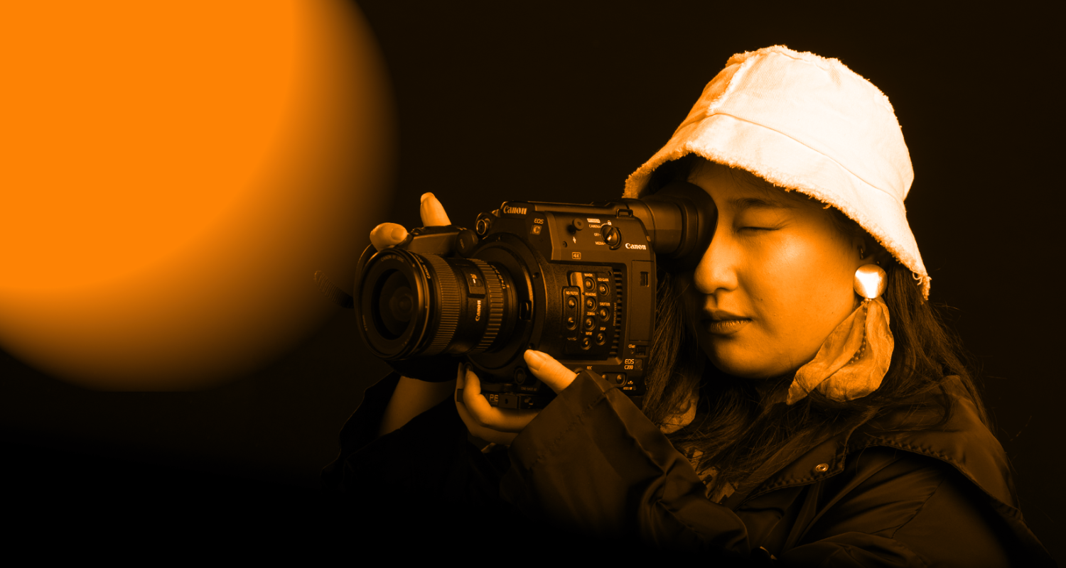 student wearing bucket hat and black jacket hold camera viewfinder up to eye focusing on something off frame with an orange wash overlay 