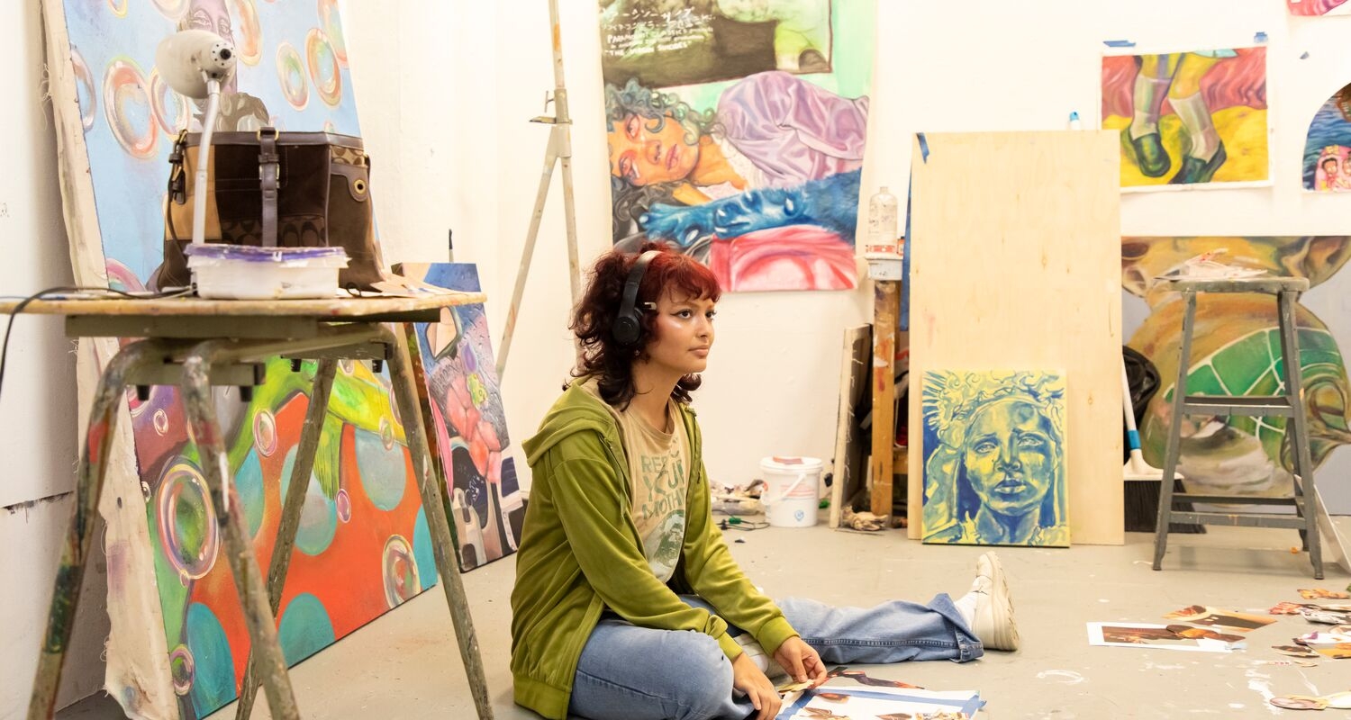 An artist sitting on the floor of their studio surround by paintings