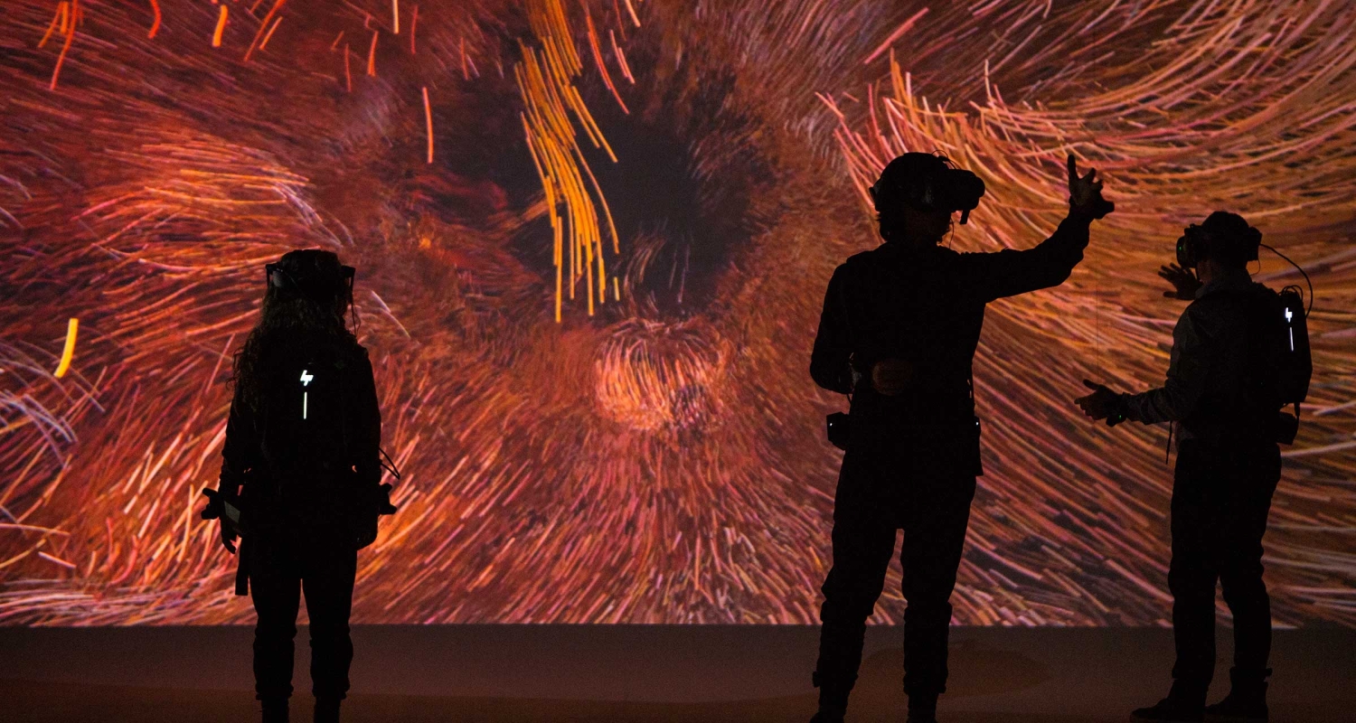 Three silhouetted students stand in front of a screen. The students are wearing VR gear. The screen is displaying a still from what appears to be an abstract, interactive art piece made up of swirling fibers in shades of red and gold. The room is very dark.