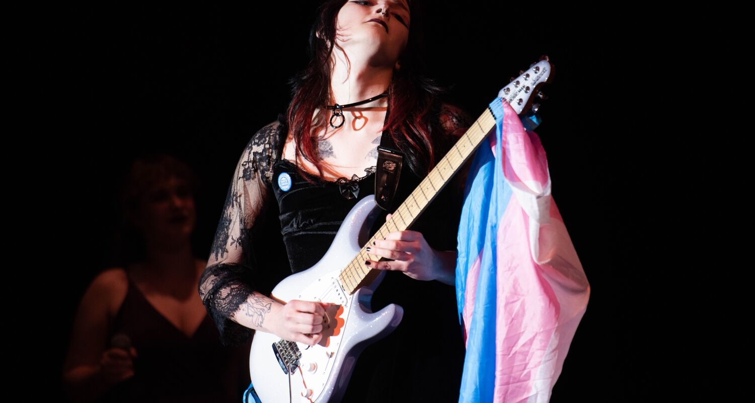A guitarist in a black dress playing an electric guitar with a trans flag hanging from the neck of the instrument