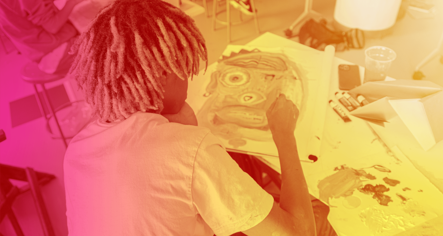 A student is working on a drawing, the viewer is looking at the student's head and shoulders. They have black and blonde streaked dreadlocks and a white teeshirt. The photograph has a gradient from pink on the left to gold on the right.