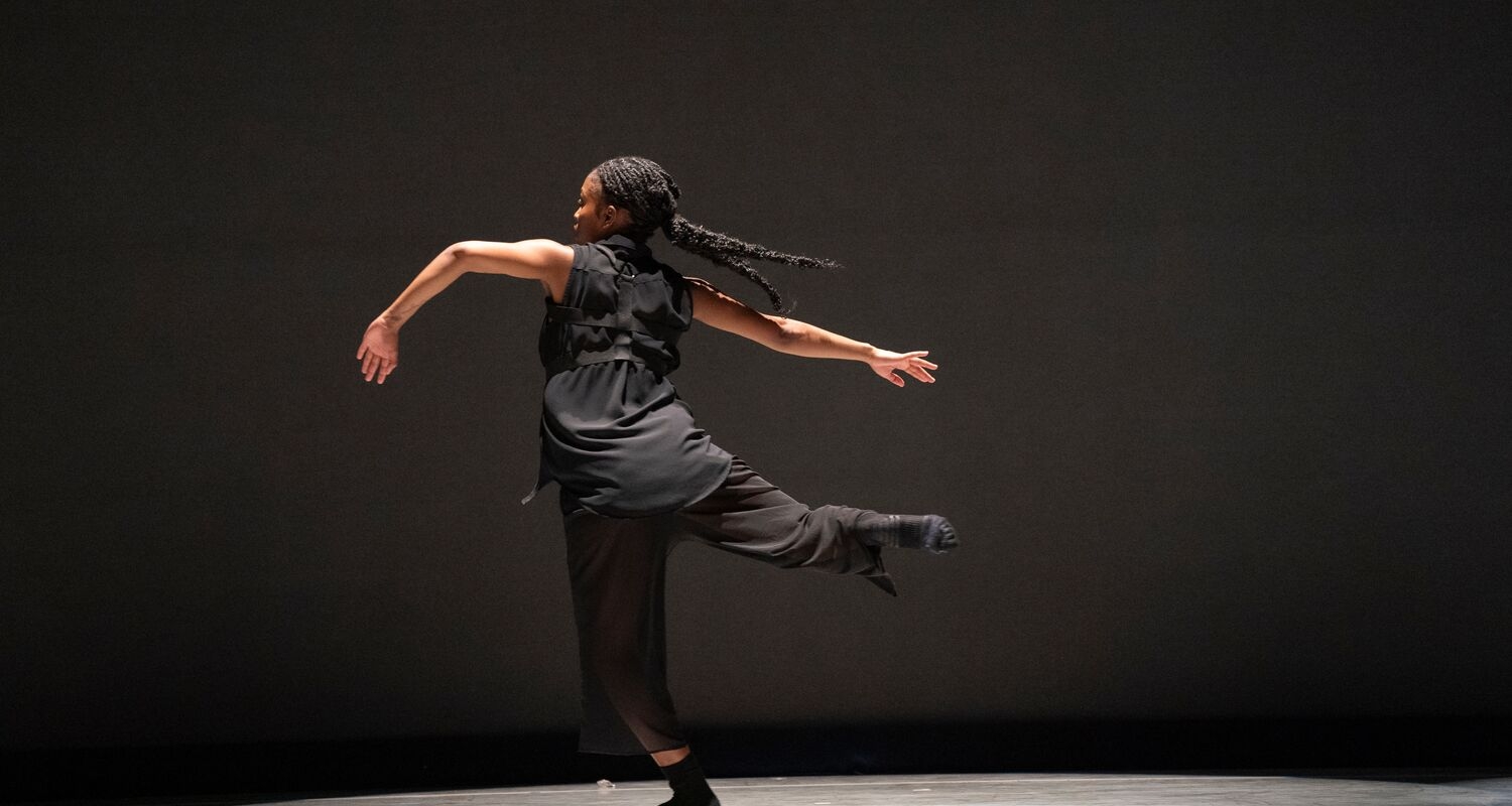 a person in a black loose outfit is dancing with back turned and arms stretched outwards against a grey-brown background 