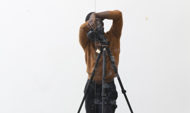 Self portrait of the artist Paul Mpagi Sepuya, artist posing in a white space behind a photo tripod with face obscured by camera