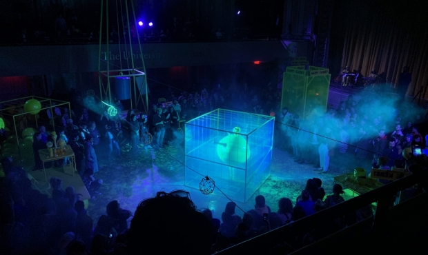 Alex Da Corte's reinvention of Alan Kaprow's Chicken performance. The photo was taken in UArts' Gershman Hall and shows a large crowd and performers in a dark exhibition space with sections illuminated with blue lights and various on-going performances including a person in a bright yellow costume inside a glowing cage.