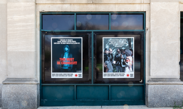 posters from the Resist Covid Take 6 exhibition installed on the windows of the Arts Bank. The poster on the left includes a single figure with the words This must be changed in large red text in the center. The other poster includes a group of people underneath a block of white text not legible in the image 