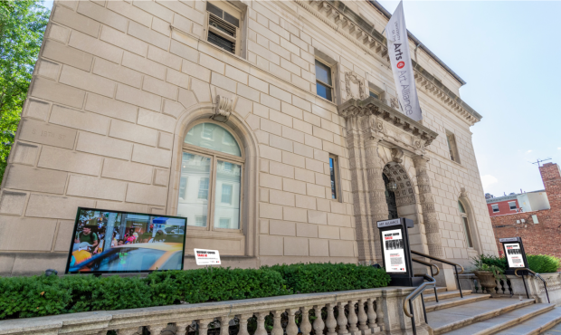 Art Alliance exterior with a digital display screen installed in the foreground of the image on the left. A white banner reads Art Alliance at the University of the Arts in black text with a red UArts logo mark. 