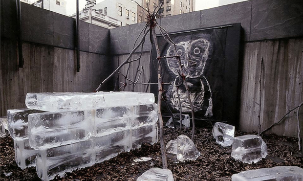 installation photograph of a work by artist Rafael Ferrer featuring large blocks of ice stacked to make a house-like structure, another structure made of branches and a drawing in white crayon on the back wall picturing a large bird. 