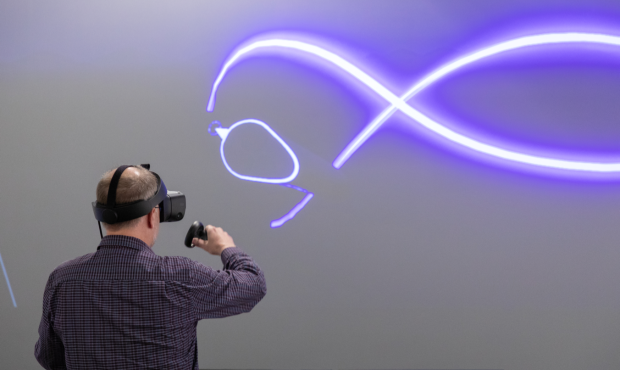 a view from behind of a person wearing a headset and holding other digital equipment facing a screen with a glowing purple line drawing on it