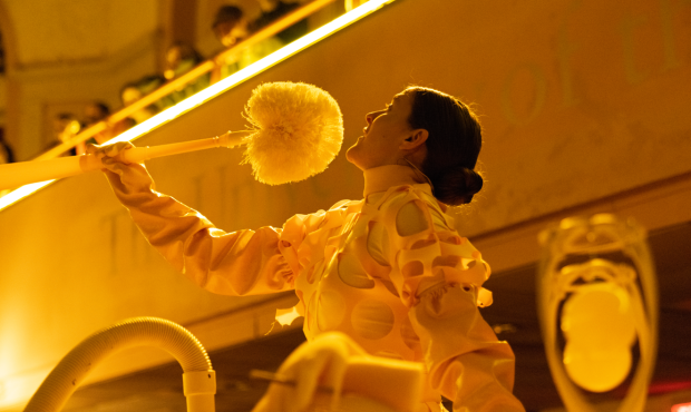 Still image from Allan Kaprow's Chicken Reimagined by Alex Da Corte depicting a performer dressed in all yellow and holding a yellow item in an auditorium, illuminated with yellow lighting