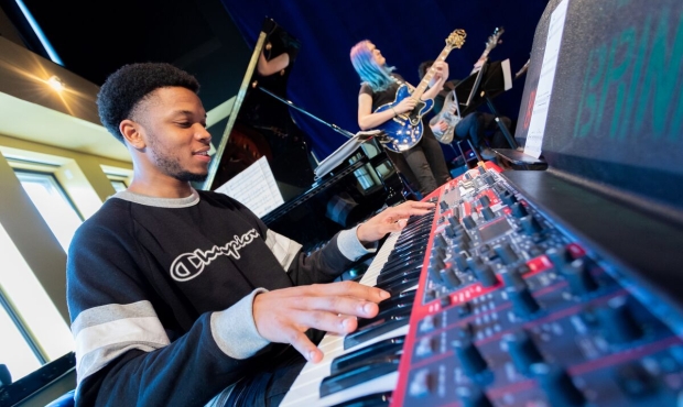 a low angle image of a person playing an electric keyboard, taken from the level of the keyboard. in the background are two people playing guitars