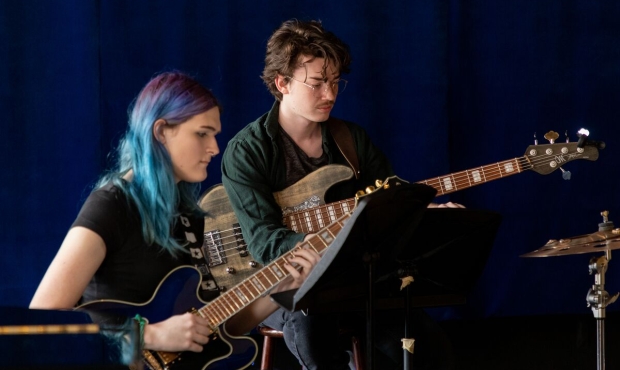 two people play guitars against a background of a blue curtain. the person in front has long teal hair, the person in the back has round glasses and a mustache. both are looking downward toward the right of the image.