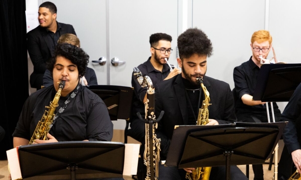 five seated people wearing black, all playing saxophones. another person stands along the wall at the back of the room wearing a black suit.