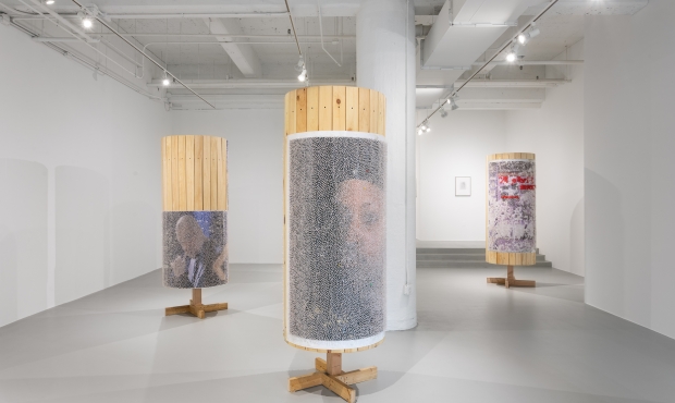 Installation view of exhibition showing three pillars made of plywood, one in foreground, two in background, covered in large posters and tacks
