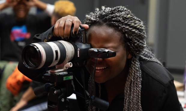 A person in a long sleeved black shirt with long gray shaded braids leans into a DSLR camera on a tripod and looks into the rangefinder with a big smile. They are gripping the camera from its grip, which is positioned at the top with the camera’s current orientation. They have black nail polish and a silver ring on their middle finger. Two other people are seen out of focus in the back, seated and speaking. 