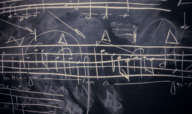 Chalk drawing of musical notation on a blackboard with smudgy poorly erased notation and musical theory notes layered below. The chalk has a yellowish shade. 
