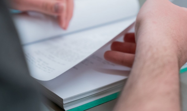 A narrow focus photo of two hands handling a screenplay book that is laid on a table. The left hand is holding down the open left half of the book as the right hand scoops up the open page and is turning it from below. 