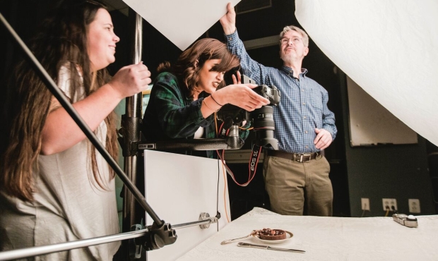 Three people are working in a photo studio to establish google lighting and take an overhead shot of a tarte on a plate. The person on the left is wearing a gray dress and is holding a lighting stand. The person in the center has a green flannel shirt and is holding a digital camera on a stand pointed directly downward at the tiny plate and silverware. the person on the left has glass and gray hair and a tucked in shirt and is adjusting an overhead lighting reflector. The tarte is on a white tablecloth cont