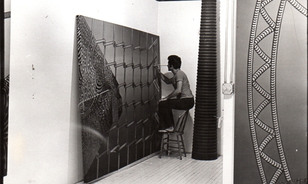 Black and white photo of man sitting on back of a chair painting  a large canvas in a studio