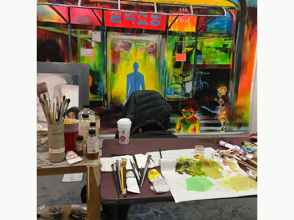 A student's painting studio. Paints, brushes, paper towels and other supplies are laid across the table with a canvas hanging on the wall behind.