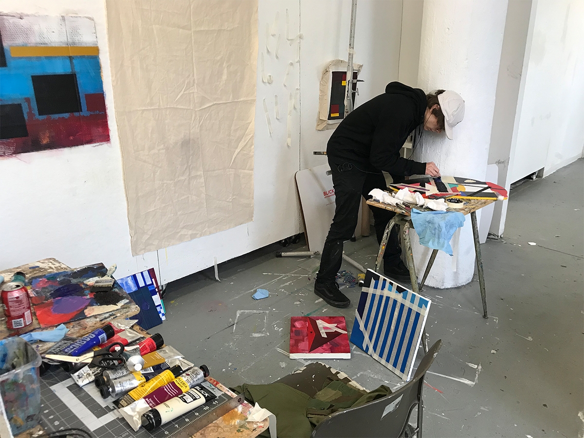 A student works in their painting studio on a piece of art. Paints are laid out across the table and canvases are propped up against the wall.