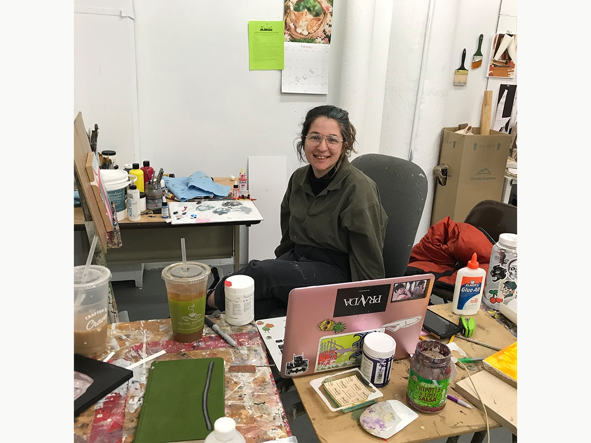 A student sits in their painting studio behind their computer and smiles at the camera.