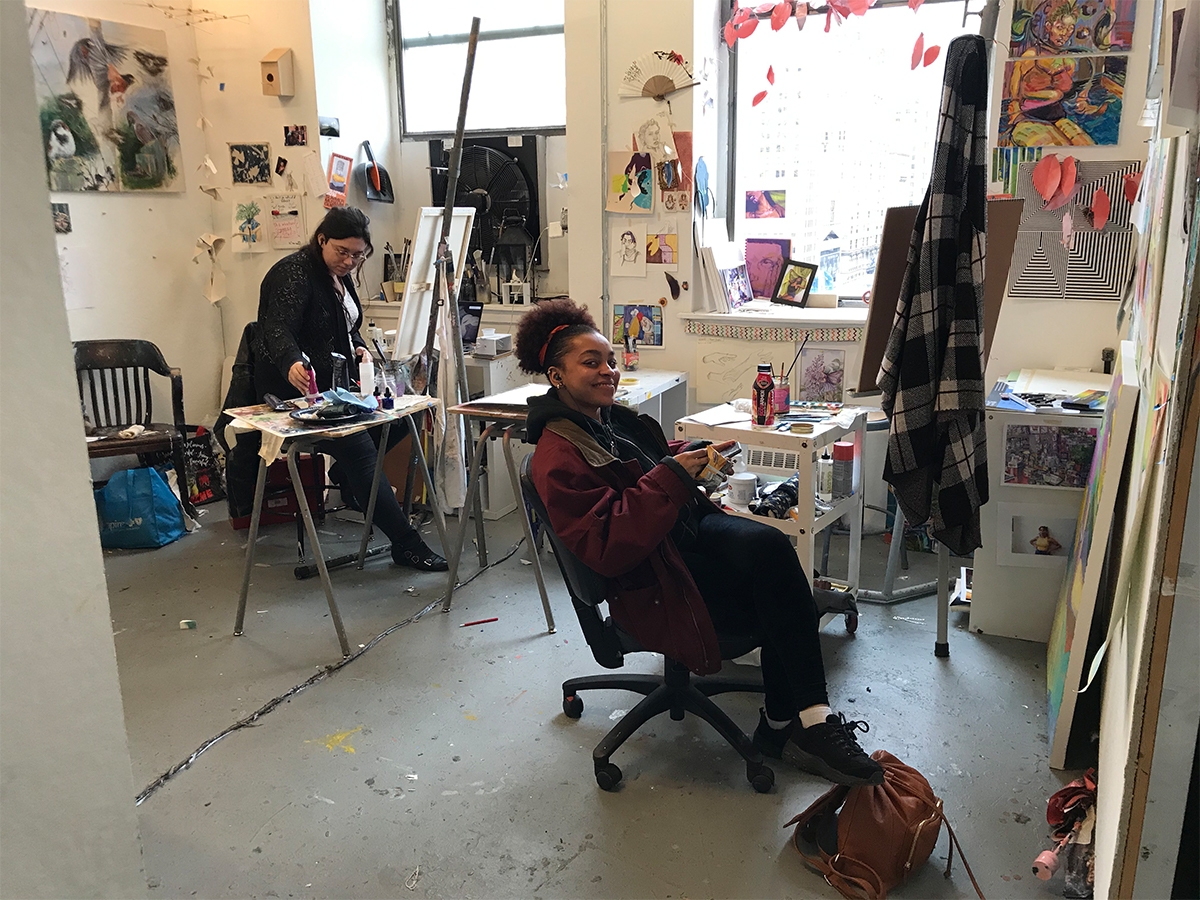 Two students work on their art in the painting studios. One student smiles at the camera while they work.