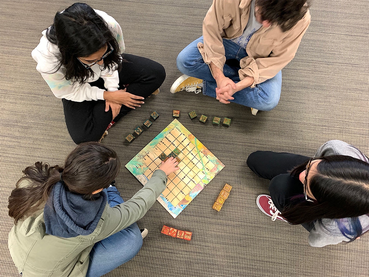 Students sit around a game board and play a game with small square wooden pieces.