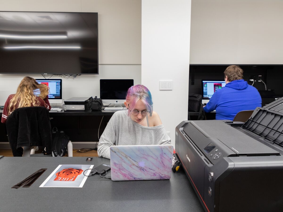 A student works on their laptop next to a printer in the digital imaging lab.
