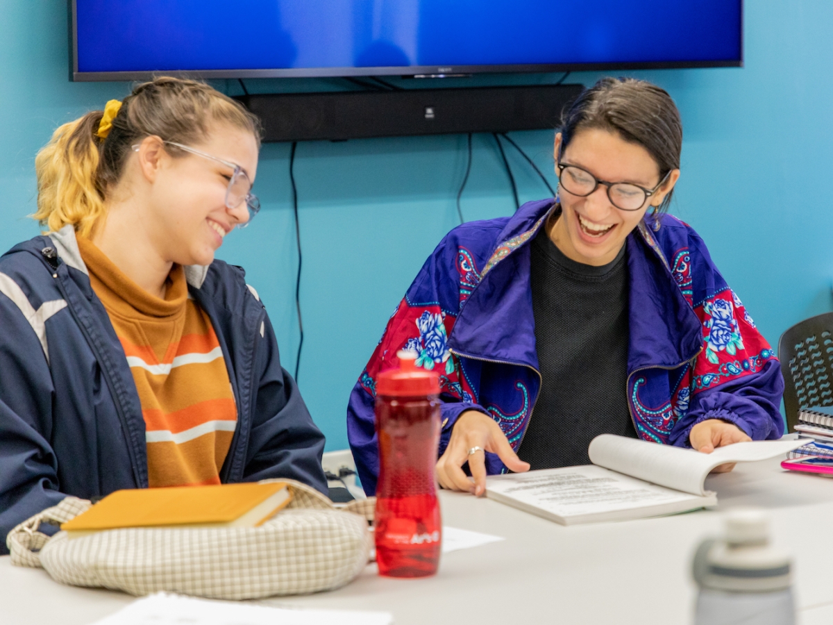 Two students sit at a table and laugh while reading a script during a Screenwriting class