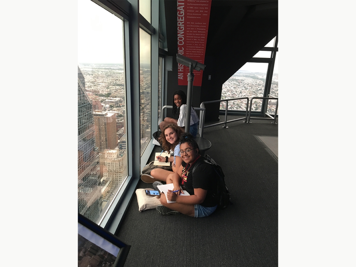 Students sit and write while looking at the Philadelphia skyline