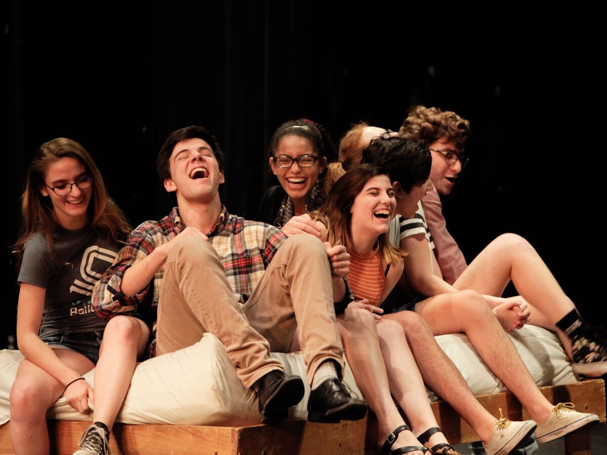 Theater students sit on a bed on stage and laugh during a performance