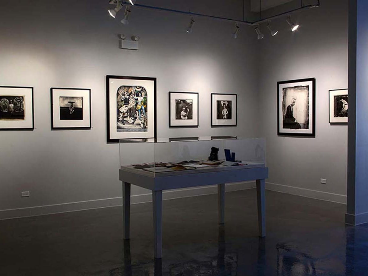 Installation view of Joel-peter Witkin: From The Studio at Catherine Edelman Gallery. Several photographs are pictured hanging on the white, gallery walls; a table in the foreground has objects under a plexiglass case.
