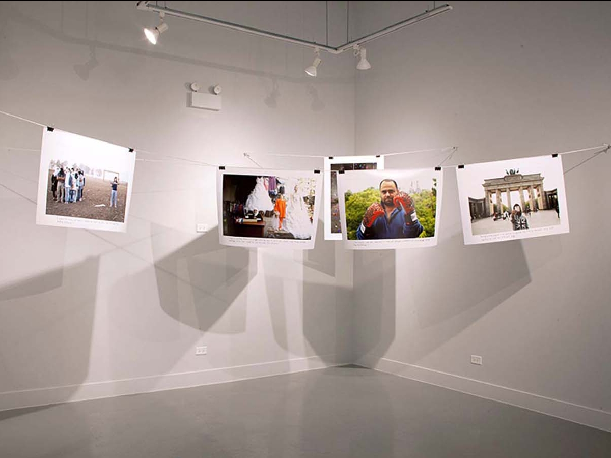 Installation view of What is Home? At Catherine Edelman Gallery. Several photographs are suspended on a wire secured by binder clips. The artwork is casting a set of grayscale shadows against the gallery wall.